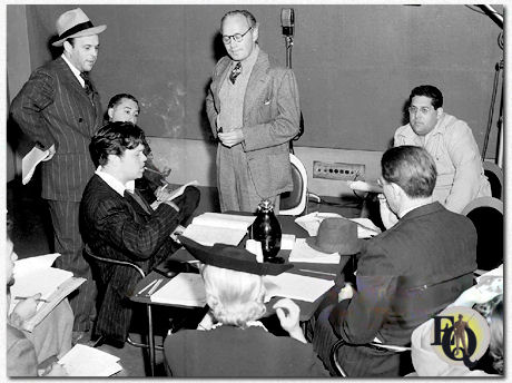 Orson Welles sits at a table, smoking his pipe, with his star, comedian Jack Benny, standing, center, and others during rehearsal of "June Moon" at the KNX radio station for the CBS Campbell Playhouse broadcast, Hollywood, California, March 24. 1940. From left: Bill Morrow (standing); Welles; Benny Rubin sitting behind Welles); Benny; Ernest Chappell (seated at table across from Welles); Jack Beloin (sitting facing camera, another Benny writer). 